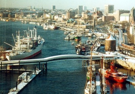 Hamburger Hafen im Schnee im Januar 2026: Blick über die zugefrorene oder schneebedeckte Elbe mit einer geschwungenen weißen Brücke im Vordergrund, mehreren großen Frachtschiffen, kleineren Booten und der Skyline der Hamburger Innenstadt im Hintergrund unter winterlichem Himmel.